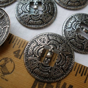 May include: Silver metal buttons with an intricate geometric design and a rectangular opening in the center. The buttons are approximately 1 inch in diameter.