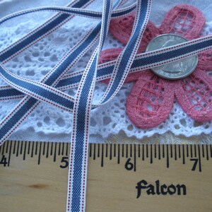 May include: A close-up of a white ribbon with blue and red stripes. The ribbon is measured with a wooden ruler. A pink flower and a coin are also in the image. The word "Falcon" is printed on the ruler.