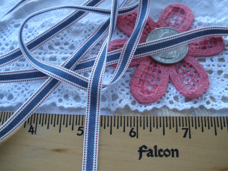 May include: A close-up of a white ribbon with blue and red stripes. The ribbon is measured with a wooden ruler. A pink flower and a coin are also in the image. The word "Falcon" is printed on the ruler.