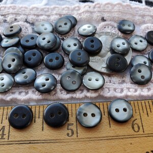 May include: A collection of dark gray and black buttons with two holes each, scattered on a lace surface. A silver coin is also present. A wooden ruler with inch markings is in the foreground, with the numbers 4, 5, and 6 visible.