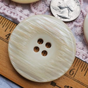 May include: A large, round, cream-colored button with four holes. The button has a swirled, pearlescent effect. A wooden ruler with inch markings and a US quarter are partially visible in the background.