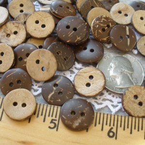 May include: A collection of brown coconut shell buttons with two holes each. The buttons are scattered on a white lace fabric with a wooden ruler in the foreground. The ruler has markings in inches.