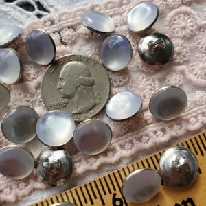May include: A collection of 14 iridescent white buttons with silver metal shanks. The buttons are arranged on a white lace fabric background with a US quarter for scale.