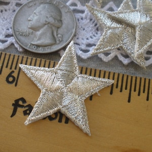 May include: Silver embroidered star patches. The patches are on a wooden ruler with inch markings. A US quarter coin is also in the image. The patches are likely for crafts or embellishments.