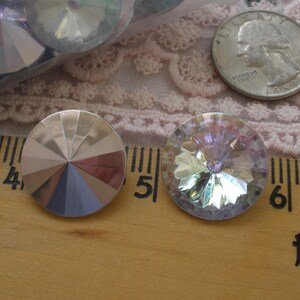 May include: Two iridescent, faceted crystal rhinestones with a quarter coin for size comparison. One rhinestone has a silver and pastel color scheme, the other has a rainbow effect. The rhinestones are on a wooden surface.