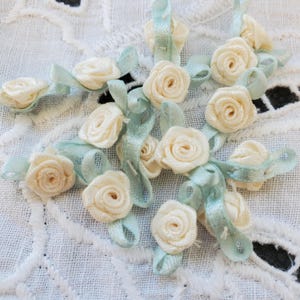 May include: Close-up of several small, cream-colored fabric roses with light blue ribbon accents. The roses are arranged on a white, textured surface, creating a delicate and floral design. The ribbons are approximately 0.5 inches wide.