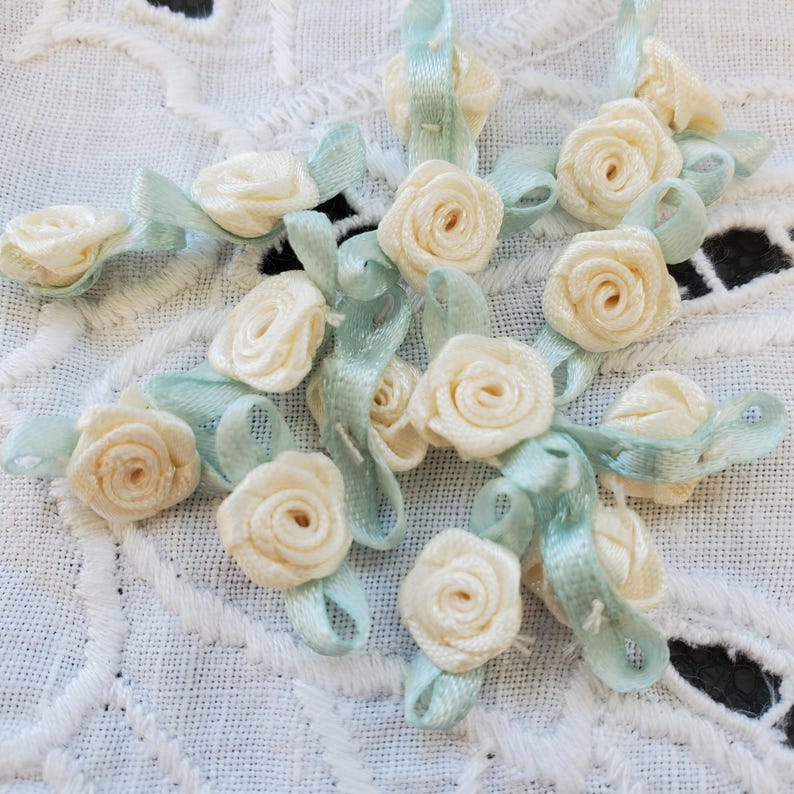 May include: Close-up of several small, cream-colored fabric roses with light blue ribbon accents. The roses are arranged on a white, textured surface, creating a delicate and floral design. The ribbons are approximately 0.5 inches wide.
