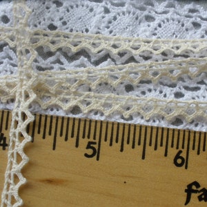 May include: Close-up of a white and cream lace trim with a scalloped edge. The trim is measured against a wooden ruler with markings in inches.