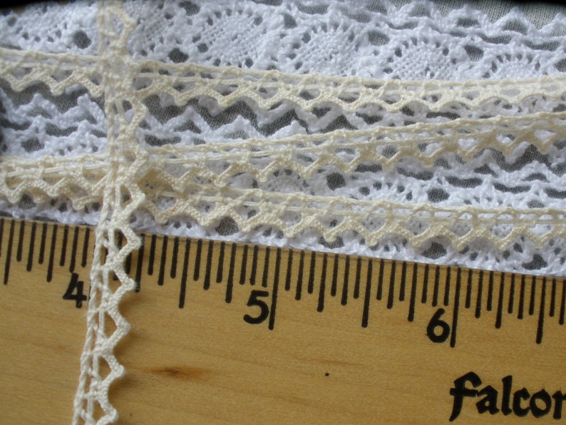 May include: Close-up of a white and cream lace trim with a scalloped edge. The trim is measured against a wooden ruler with markings in inches.