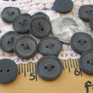May include: A collection of dark gray buttons with two holes and concentric circle detailing. A US quarter is included for size comparison. The buttons are arranged on a wooden ruler and lace fabric.