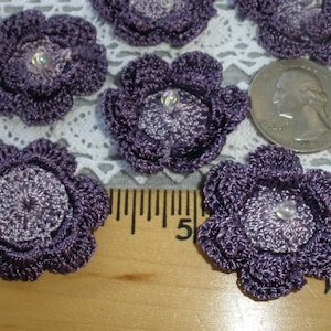 May include: Six purple crocheted flower embellishments with a pearl-like center. The flowers are approximately the size of a US quarter.