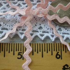 May include: Pink zig zag trim with a white crochet lace background. The trim is measured on a ruler with the numbers 5 and 6 visible.