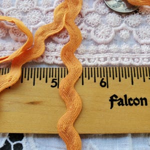 May include: Close-up of orange wavy ribbon with a textured surface. The ribbon is next to a wooden ruler with black markings and the word "Falcon" printed on it. The background includes a lace fabric and a coin.