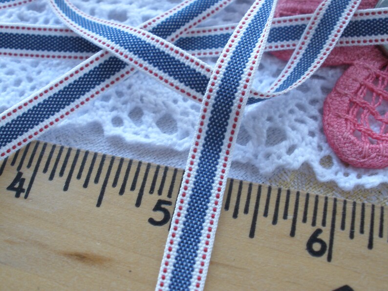 May include: Close-up of a blue, white, and red striped ribbon. The ribbon is approximately 0.5 inches wide and features a repeating pattern of blue stripes, white stripes, and red dots. A wooden ruler with inch markings is in the foreground.