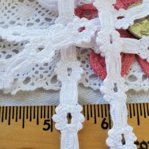May include: White cotton lace trim with a delicate floral design. The trim is approximately 1/2 inch wide and features a repeating pattern of small flowers with open centers. The lace is shown against a ruler, with the measurement markings visible.