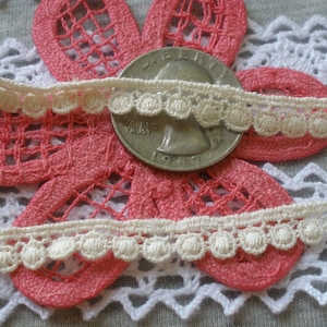 May include: Two pieces of white crocheted trim with a scalloped edge and small round embellishments. The trim is shown on top of a pink and white crocheted doily.