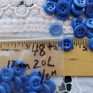 May include: A collection of small, round, blue plastic buttons with four holes in the center. The buttons are stored in a clear plastic bag marked with handwritten text including 13mm, 20L, and 1/2 inch, positioned next to a wooden ruler.