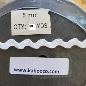 May include: White, wavy-edged ribbon, 5 mm wide, on a spool. The label reads "QTY: 20 YDS" and "www.kaboooco.com". The ribbon is likely used for sewing, crafting, or decorative purposes.
