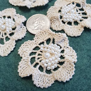 May include: Close-up of several cream-colored, crocheted flower appliques with pearl bead centers. A U.S. quarter coin is next to the appliques for size comparison. The appliques are on a dark green surface.