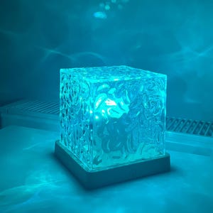 Puede incluir: Una lámpara cuadrada de vidrio transparente texturizado con un brillo azul. La lámpara se asienta sobre una base blanca y emite una luz ambiental suave. El vidrio tiene un patrón orgánico detallado. El fondo es un degradado de azul.