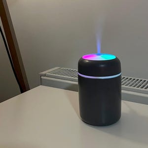 May include: A dark gray essential oil diffuser with a colorful, illuminated top. The top of the diffuser has a ring of light and emits a fine mist. The diffuser sits on a white surface.