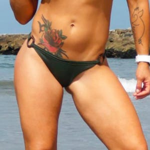 May include: A person wearing a dark green bikini bottom standing in the ocean. The person has a rose tattoo on their abdomen and a tattoo on their arm. A white watch is on their wrist. The ocean is in the background.