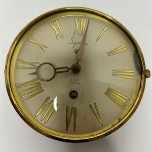 Può includere: Un quadrante di orologio rotondo e vintage con una cornice e lancette color oro. Il quadrante è bianco sporco con numeri romani dorati e il testo "Syro" e "8 Day Jeweled". Le lancette dell'orologio indicano circa le 1:07.