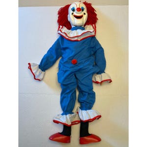 Könnte beinhalten: Eine Bauchredner-Clownpuppe mit weißem Gesicht, blauen Augen, roter Nase und breitem Lächeln. Die Puppe hat leuchtend rotes Haar, einen blauen Overall mit rotem Bommel, einen weißen Rüschenkragen und Manschetten mit rotem Saum sowie schwarze Leggings und rote Schuhe.