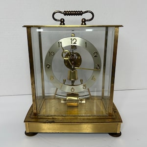 Può includere: Un orologio rettangolare con cornice in ottone e cassa in vetro trasparente. Il quadrante è argentato con numeri e lancette nere. C'è una maniglia decorativa in alto. L'orologio ha un pendolo e una campana.