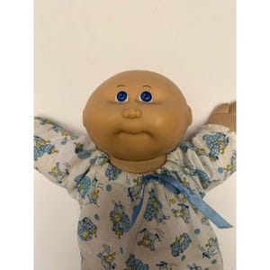 Puede incluir: Muñeca Cabbage Patch Kid vintage con cabeza calva y ojos azules. La muñeca lleva un pelele blanco y azul estampado con una cinta azul. Los brazos de la muñeca están extendidos.