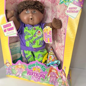 Pode incluir: Boneca Cabbage Patch Kids Birthday Kids na caixa original. A boneca tem cabelo castanho e veste uma roupa roxa e verde. A caixa inclui um chapéu de festa, uma certidão de nascimento e uma etiqueta com "Happy Birthday".