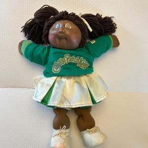 May include: A Cabbage Patch Kids doll with dark skin, brown yarn hair in pigtails, and blue eyes. The doll wears a green and white cheerleader outfit with the words "Cabbage Patch Kids" on the shirt.