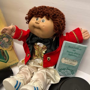 Puede incluir: Muñeca Cabbage Patch Kids, vestida con una chaqueta roja con ribetes dorados, una pajarita negra y pantalones de satén blanco. La muñeca tiene cabello castaño rizado y sostiene un certificado de nacimiento. Juguete vintage.