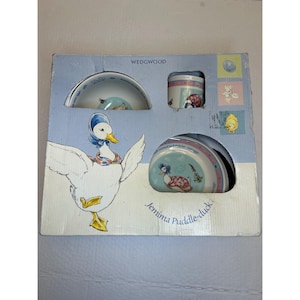 May include: A Wedgwood Jemima Puddle-duck ceramic dinnerware set in a light blue and cream cardboard box. The set includes a bowl, a mug, and a plate, each featuring illustrations of the duck character. The box displays the name Jemima Puddle-duck.