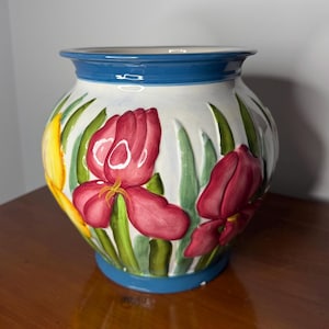 May include: A ceramic vase with a white base, adorned with red and yellow iris flowers and green leaves. The rim and base are painted blue, creating a vibrant contrast. The vase is a decorative piece.