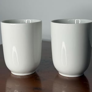 May include: Two white ceramic cups with a smooth, rounded design. The cups are cylindrical with a slightly flared rim and sit on a dark wooden surface. The cups are empty and have a minimalist aesthetic.