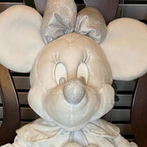 Puede incluir: Una mu&ntilde;eca de peluche de Minnie Mouse con grandes orejas redondas y un lazo plateado. La mu&ntilde;eca tiene una cara blanca, una nariz plateada y pesta&ntilde;as bordadas. Lleva una capa blanca con detalles de copos de nieve.