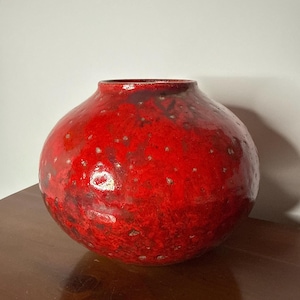 May include: A round, red ceramic vase with a textured surface and small white speckles. The vase has a wide body and a narrow opening at the top. The vase is sitting on a wooden surface.
