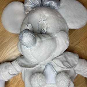 Puede incluir: Mu&ntilde;eca de peluche de Minnie Mouse con un atuendo de invierno. La mu&ntilde;eca es principalmente blanca con detalles plateados, incluyendo un lazo, nariz y detalles de copos de nieve en el vestido. Tiene orejas grandes y redondas y rasgos faciales bordados.