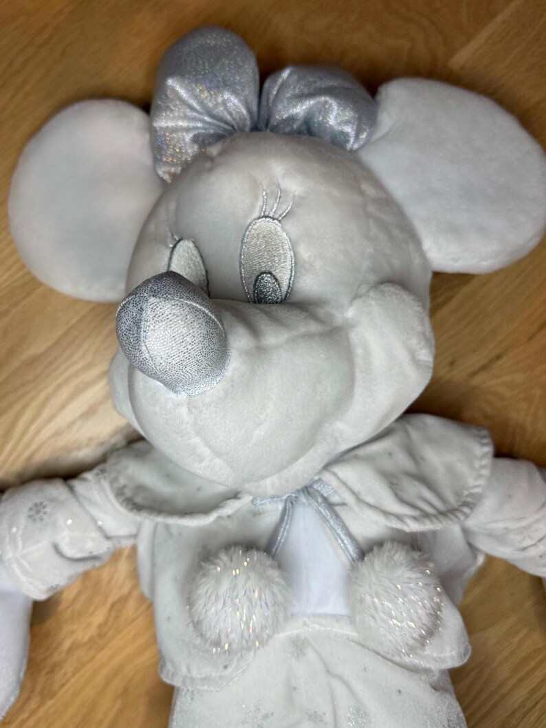Puede incluir: Mu&ntilde;eca de peluche de Minnie Mouse con un atuendo de invierno. La mu&ntilde;eca es principalmente blanca con detalles plateados, incluyendo un lazo, nariz y detalles de copos de nieve en el vestido. Tiene orejas grandes y redondas y rasgos faciales bordados.