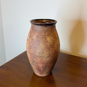May include: A textured, brown vase with a dark brown rim. The vase has a wide body that tapers towards the base and a slightly flared opening. The surface has a mottled appearance with shades of brown and tan.