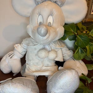Puede incluir: Una mu&ntilde;eca de peluche de Minnie Mouse con un atuendo de invierno. La mu&ntilde;eca tiene grandes orejas blancas, un lazo plateado y una nariz plateada. Su vestido es blanco con detalles de copos de nieve y dos pompones blancos. Los zapatos y guantes de la mu&ntilde;eca son plateados.