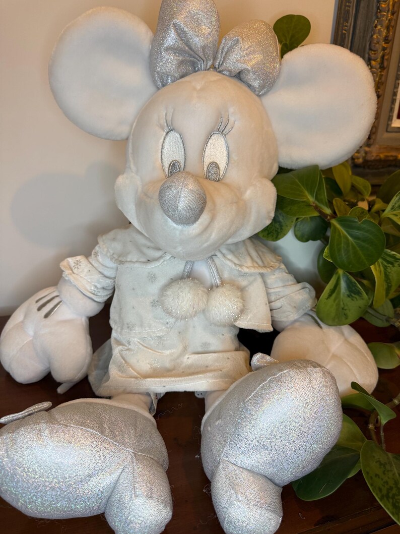 Puede incluir: Una mu&ntilde;eca de peluche de Minnie Mouse con un atuendo de invierno. La mu&ntilde;eca tiene grandes orejas blancas, un lazo plateado y una nariz plateada. Su vestido es blanco con detalles de copos de nieve y dos pompones blancos. Los zapatos y guantes de la mu&ntilde;eca son plateados.