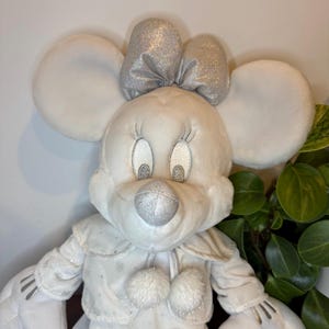 Puede incluir: Un peluche blanco con forma de rat&oacute;n con grandes orejas redondas y un lazo plateado. El rat&oacute;n tiene ojos bordados y una nariz plateada. Lleva un abrigo blanco con detalles de copos de nieve y dos pompones. Un art&iacute;culo decorativo.