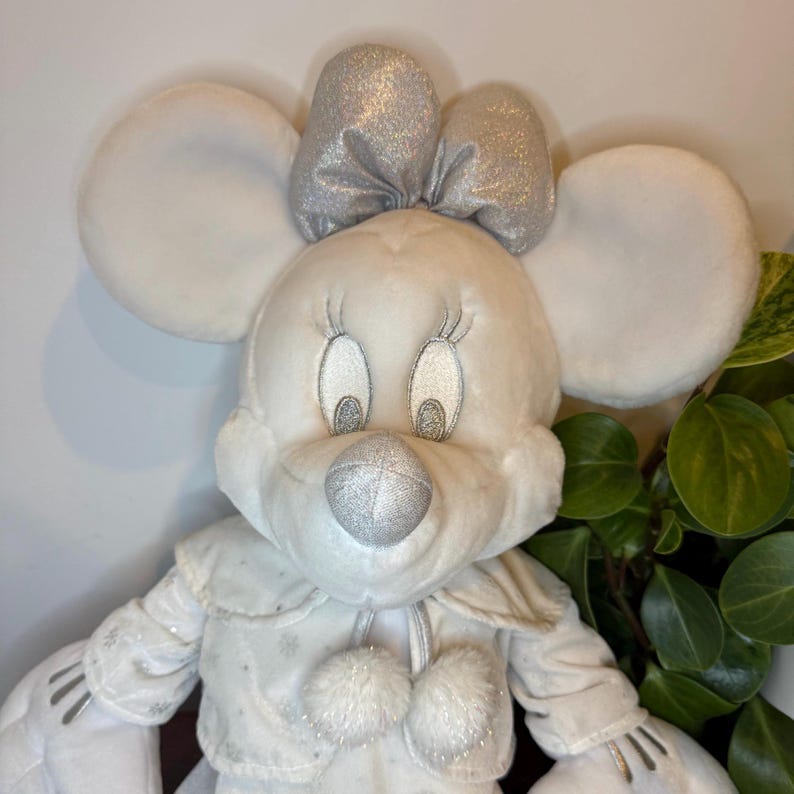 Puede incluir: Un peluche blanco con forma de rat&oacute;n con grandes orejas redondas y un lazo plateado. El rat&oacute;n tiene ojos bordados y una nariz plateada. Lleva un abrigo blanco con detalles de copos de nieve y dos pompones. Un art&iacute;culo decorativo.