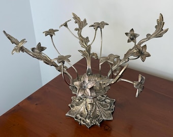 May include: Silver-toned metal floral centerpiece. The decorative piece features a base with leaf details, supporting multiple stems with flower and leaf accents. The design is intricate and detailed, suitable for home decor.