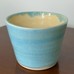 May include: A handmade ceramic cup with a light blue exterior and a tan interior. The cup has a slightly textured surface and a rounded shape. The rim of the cup has a darker blue accent. The cup is sitting on a wooden surface.