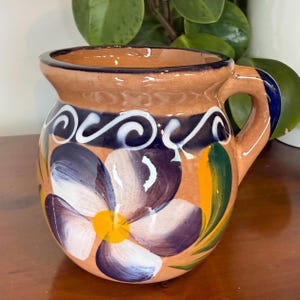 May include: Hand-painted ceramic pitcher with a floral design. The pitcher features a tan base with a dark blue band and white swirls. A large purple and white flower with a yellow center is painted on the front.