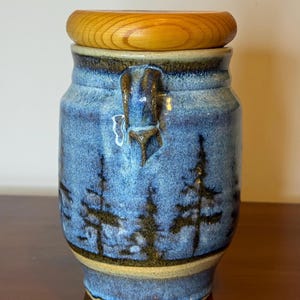 May include: A blue ceramic jar with a wooden lid. The jar features a forest scene with dark trees. The jar has a handle and a light brown base. The lid is a light brown color.