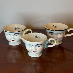 May include: Three cream-colored ceramic mugs and a creamer with whimsical faces. Each piece has a smiling face with blue eyes, a red mouth, and the word "Yum" inside. The creamer has a spout and a green bow on the handle.